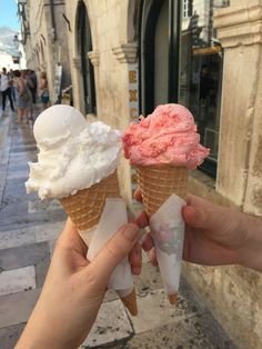 two ice cream cones with pink and white toppings in front of a stone building