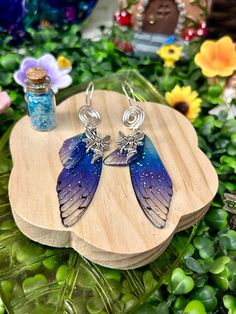 two blue and purple earrings sitting on top of a wooden board next to flowers in a vase