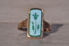 The Lynnfield: Antique Floral Carved Cameo Ring in Yellow Gold. This striking ring showcases an elongated tablet of chalcedony, adorned with a delicately hand-engraved flower motif in white and green hues, securely set within a flush bezel. On either side of the center, intricate hand-engraved patterns adorn the top of the shank, adding a touch of intricate detail to the design. Crafted with care in yellow gold, the ring exudes warmth and elegance. Currently sized at 9.25.