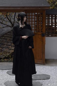 a woman dressed in black holding an umbrella and standing on gravel area next to building