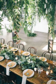 a long wooden table topped with lots of plates and place settings covered in greenery