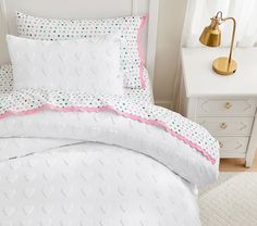 a white bed with pink and green polka dots on the comforter is next to a gold lamp