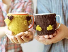 two people holding coffee mugs with knitted animals on them, one is yellow and the other is brown