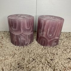 two purple candles sitting next to each other on a carpeted floor in front of a white wall