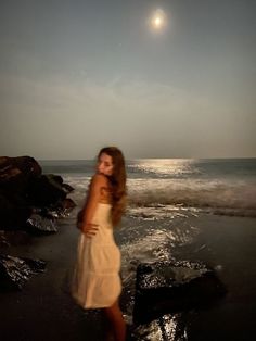 a woman standing on top of a beach next to the ocean under a full moon