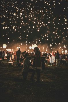 many people are standing in the grass with lanterns floating above them at night time,