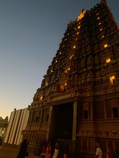 people are walking in front of an ornate building at night with lights on the top