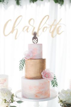 a three tiered cake with pink and gold frosting, topped with pineapple