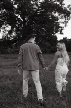 black and white photograph of man and woman holding hands walking through field with trees in the background