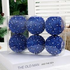 six blue balls in a clear box on top of a white table next to a christmas tree