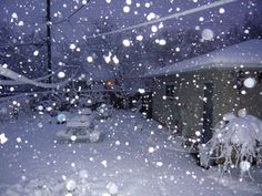 snow is falling on the street and cars are parked in front of houses at night