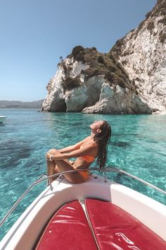 a woman sitting on the bow of a boat in clear blue water next to an island