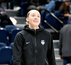 a woman standing in front of a basketball court wearing a black jacket and smiling at the camera