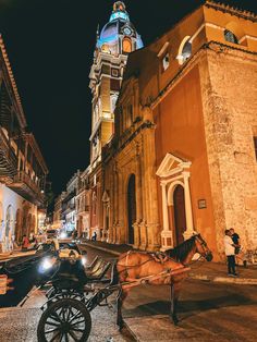 a horse drawn carriage in front of an old building at night with people standing on the sidewalk