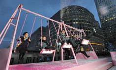 two people sitting on swings in front of a cityscape at night with buildings and skyscrapers behind them