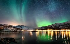 the northern lights shine brightly over water and snow covered mountains in the distance, as seen from shore