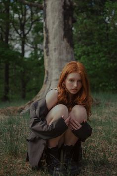 a woman with red hair sitting in the grass next to a tree and looking at the camera