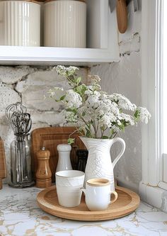 a white pitcher with flowers in it sitting on a plate next to two mugs