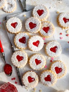 heart - shaped sugar cookies with powdered sugar and red jelly on top, next to a pair of utensils