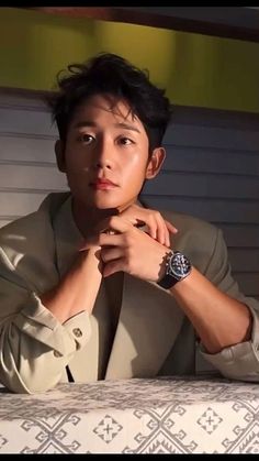 a young man sitting on top of a bed next to a white wall and wearing a watch