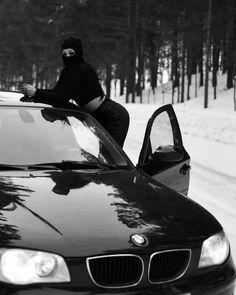 black and white photograph of a person sitting on the hood of a car in the snow