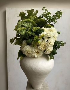 a white vase with green and white flowers in it sitting on a table next to a wall