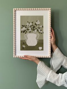 a woman holding up a framed art print with flowers in a vase on the wall