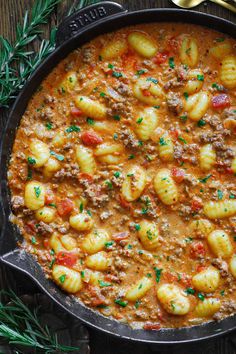 a skillet filled with pasta and sauce on top of a wooden table next to spoons