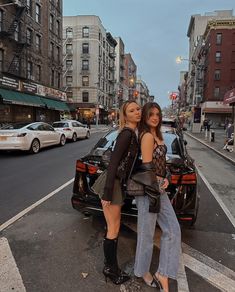 two beautiful women standing next to each other in front of a car on the street