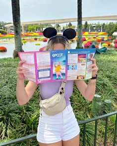 a woman in white shorts and mickey ears holding up a disneyland world book with her hands