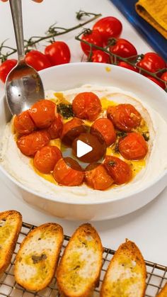 a white bowl filled with food next to toasted bread and tomatoes on a table