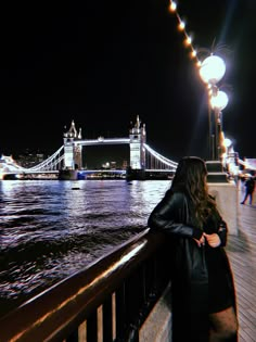 a woman standing on the side of a bridge looking at the water and lights in the background