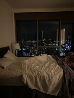 an unmade bed in front of a large window with city lights on the other side