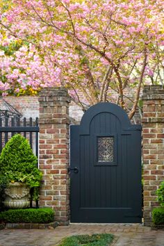 a black gate surrounded by bushes and trees with pink flowers in the background, on a brick walkway