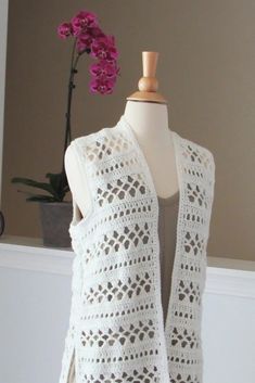 a white crocheted vest on a mannequin next to a purple flower