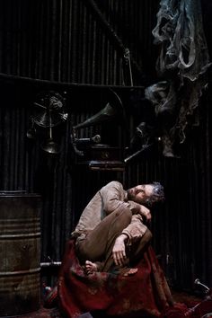 a man laying on top of a red blanket in front of a metal barrel filled with stuff