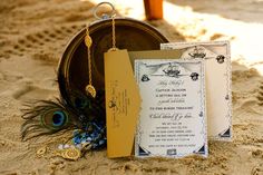 the wedding stationery is set up in the sand with peacock feathers and beads on it