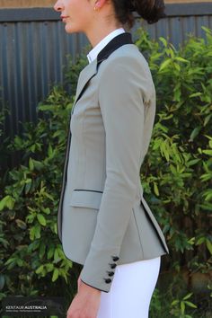 a woman wearing a gray jacket and white pants is standing in front of some bushes