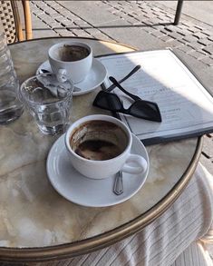 two cups of coffee sit on a table next to an open book and water glasses