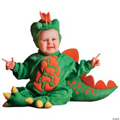 a baby in a green dragon costume sitting on the ground with his hands up and smiling