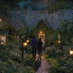 two people walking down a path in front of a house with lanterns on the walkway