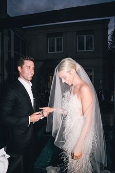 a man and woman standing next to each other in front of a wedding dress with feathers on it