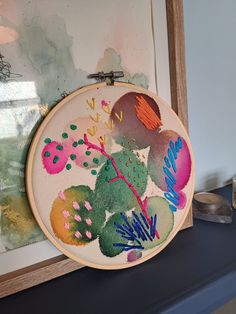 an embroidery project is being displayed on a shelf in front of a framed painting and other items