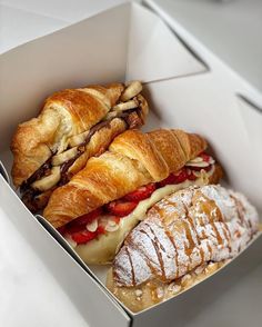 two croissant sandwiches in a box with powdered sugar on the side and pastries