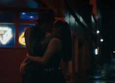 a man and woman embracing on the street in the dark with neon signs behind them