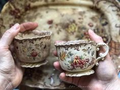 two hands are holding small cups in front of an old china plate with floral designs on it