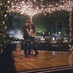 a man and woman standing on top of a wooden floor covered in christmas lights at night