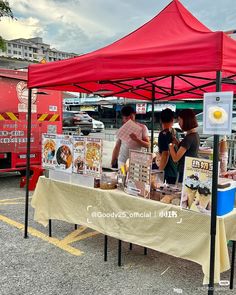 some people are standing under a red tent at an outdoor food stand with posters on it