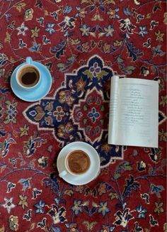 two cups of coffee sit next to an open book on top of a red rug