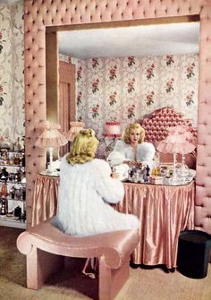 two women sitting at a vanity in a room with pink furniture and wall papered walls